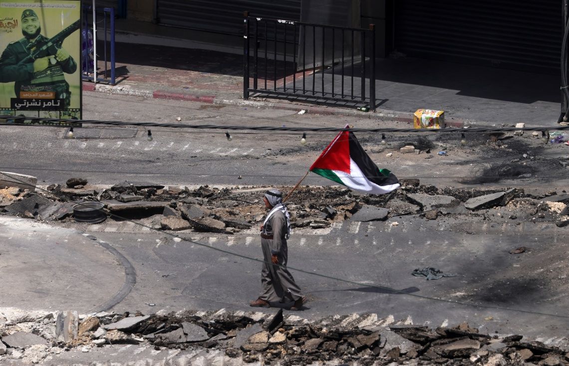 Un palestino ondea una bandera palestina mientras camina por una calle dañada durante el sexto día de una operación militar israelí en la ciudad cisjordana de Yenín, este lunes. Foto: Alaa Badarneh / EFE.